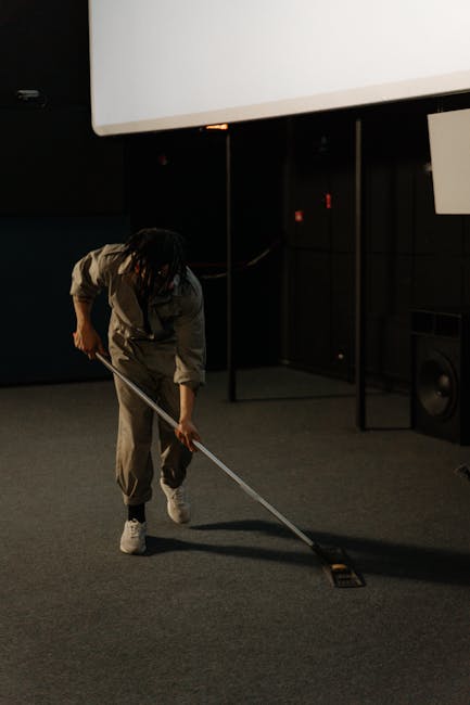 A professional cleaner dressed in white protective suit, including a hood, gloves, and blue shoe covers, is using a vacuum or steam cleaning machine on a beige carpet in a well-lit living room. The ro