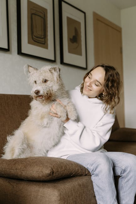 A cozy living room scene featuring a white fabric sofa with two children sitting on it. The children, a boy and a girl, are playing with a small, fluffy brown puppy. The room has a modern aesthetic wi